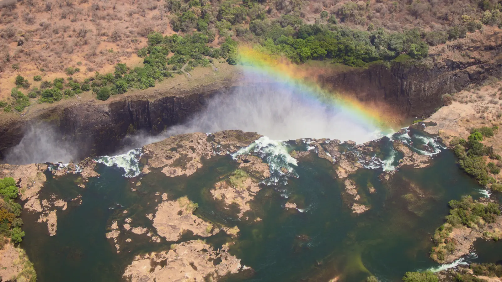 Spray and a rainbow rise from a wide waterfall dropping into a dark basalt gorge.
