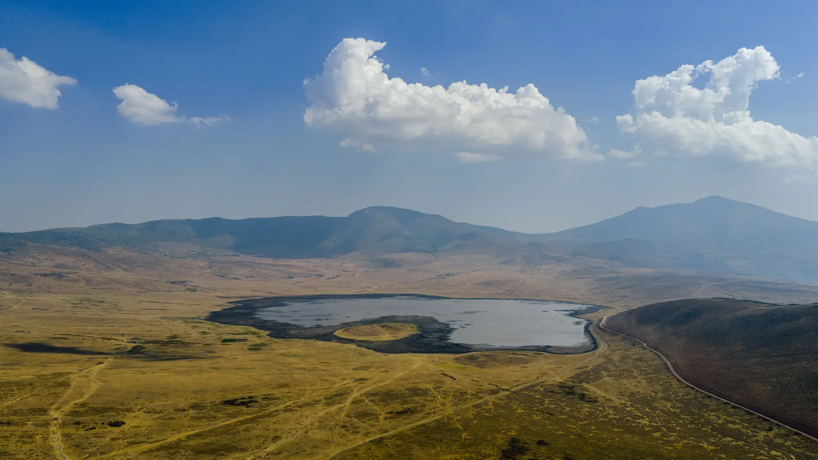A shallow soda lake sits in a broad grassy caldera ringed by green volcanic hills.