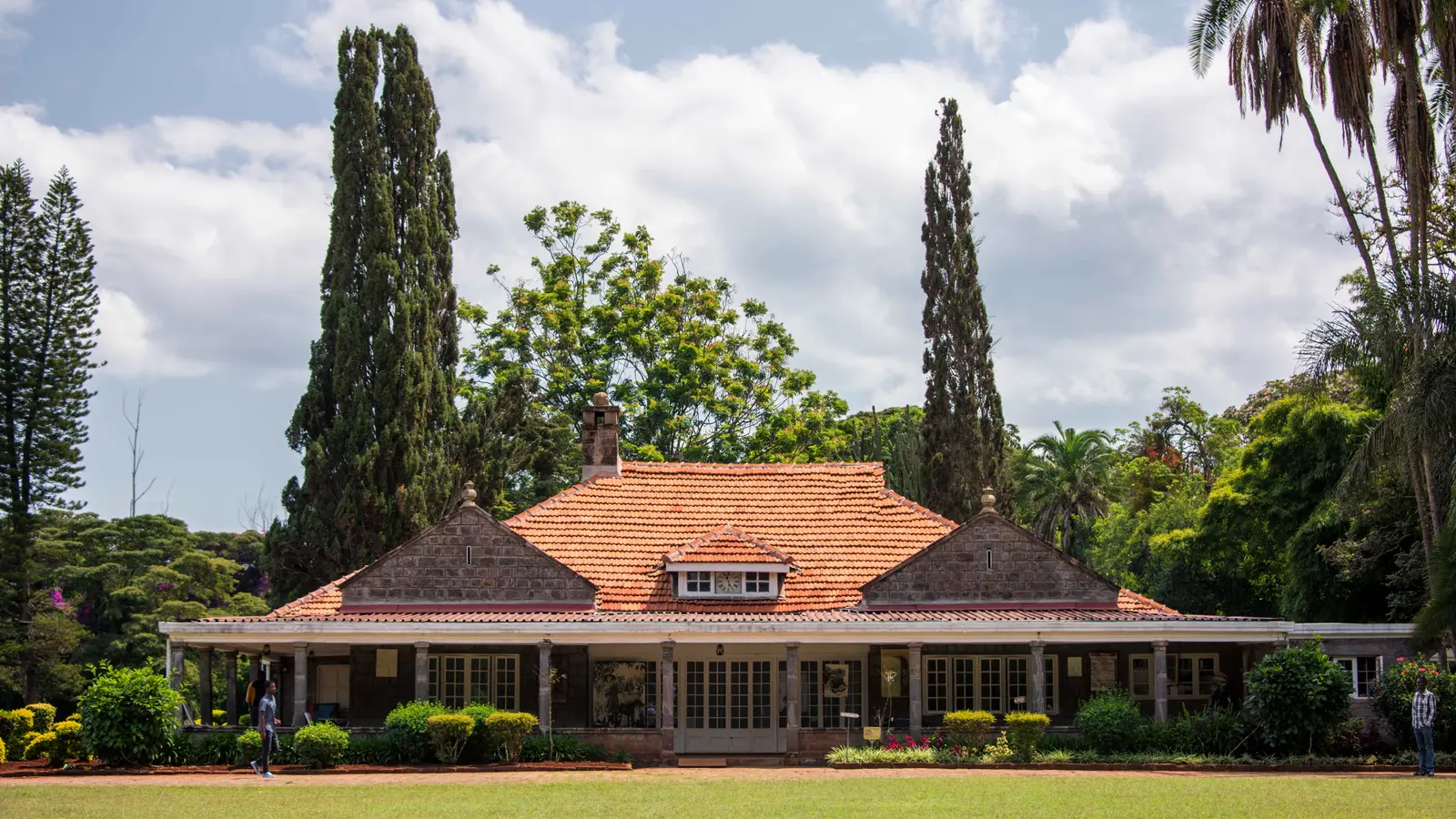 A tiled-roof stone house with a long verandah stands behind lawn and tall cypress trees.