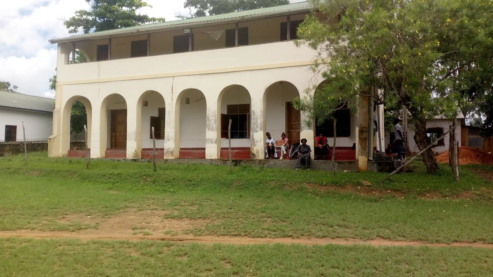 A white arcaded mission building with a green roof stands behind open grass.