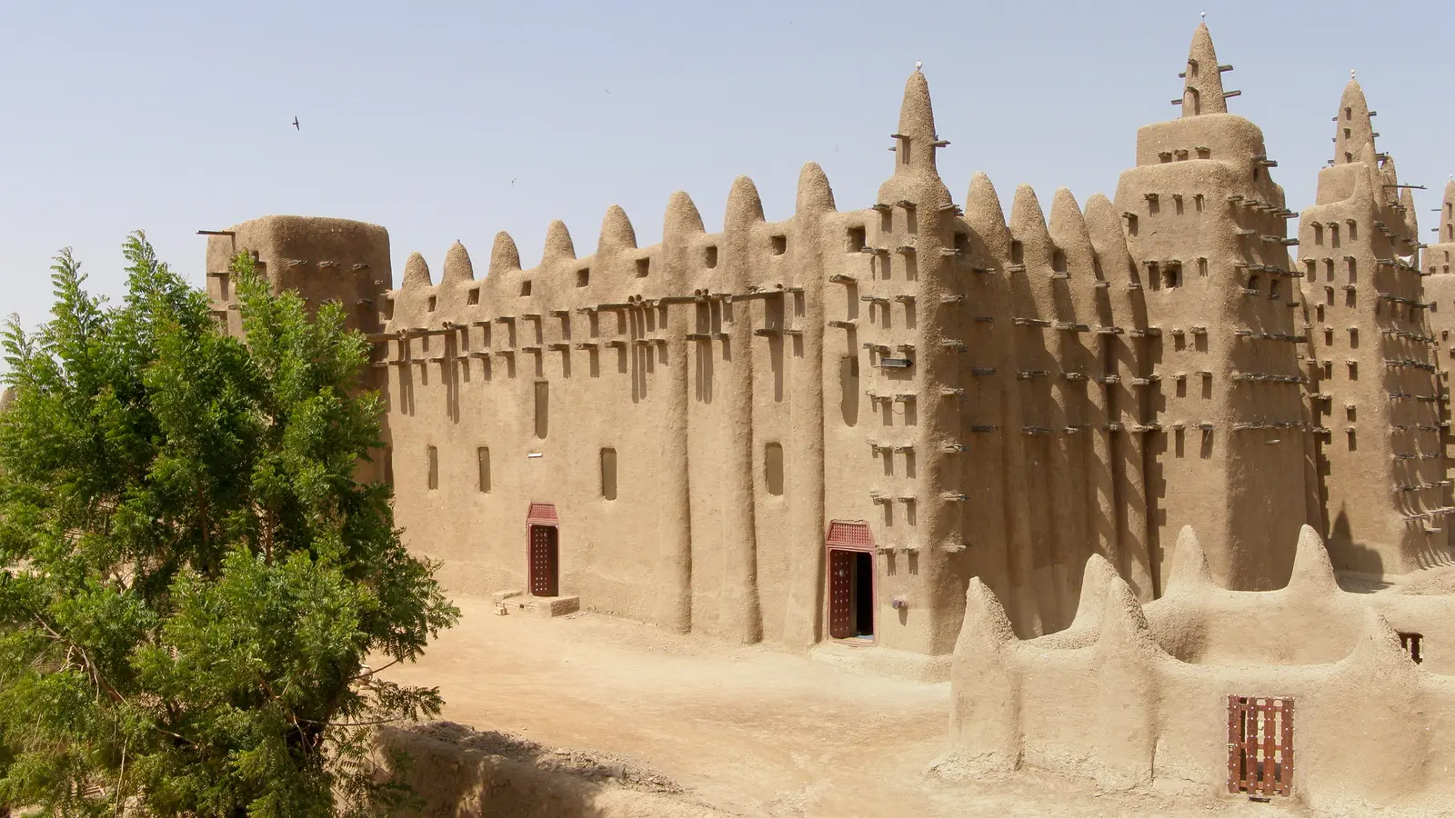 A large mud-brick mosque with tapered towers and projecting timber beams stands above bare ground.